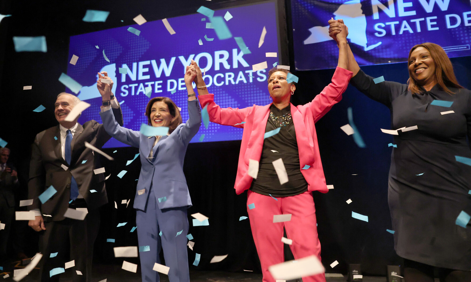 Tom DiNapoli, Kathy Hochul, Adrienne Adams, and Letitia James hold hands in the air in celebration as colorful confetti falls through the air.